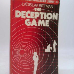 May include: A vintage book titled "The Deception Game" by Ladislav Bittman. The cover is predominantly red with white concentric circles and the title in large white letters. Silhouettes of figures are also visible.