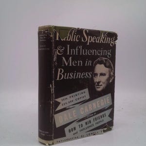 May include: A vintage hardcover book titled "Public Speaking & Influencing Men in Business" by Dale Carnegie. The cover is dark with white text and a portrait. The book shows signs of wear, with visible damage to the edges.