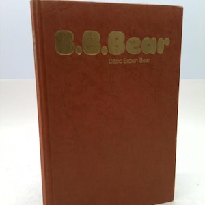 May include: A brown book titled "B.B. Bear" with gold lettering. The text "Basic Brown Bear" is below the title. The book has a textured cover and is slightly angled, showcasing its spine.