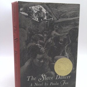 May include: A black and white illustration of a book cover for "The Slave Dancer" by Paula Fox. The cover features a drawing of a ship's hold with enslaved people packed inside. The book title is written in a stylized font with a gold seal.