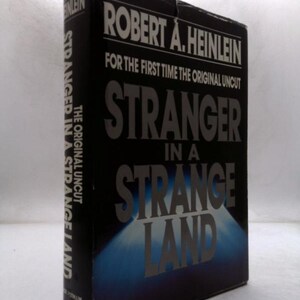 May include: A hardcover book titled "Stranger in a Strange Land" by Robert A. Heinlein. The book has a black cover with white and gray text. The title is prominently displayed, with the words "Stranger" and "Land" in large font.