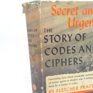 May include: A hardcover book titled Secret and Urgent: The Story of Codes and Ciphers by Fletcher Pratt. The dust jacket features a dark gray background with scattered letters and numbers, orange text, and a rectangular white information box.
