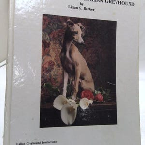 May include: A hardcover book titled The New Complete Italian Greyhound by Lilian S. Barber. The white cover features a central photograph of a tan Italian Greyhound sitting among white calla lilies and red roses against a dark floral backdrop.
