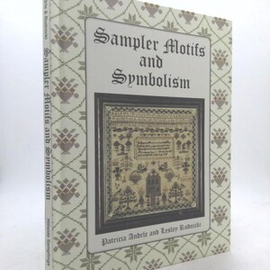 May include: A hardcover book titled "Sampler Motifs and Symbolism" by Patricia Andrle and Lesley Rudnicki. The cover features a framed sampler with a detailed design. The book's spine displays the title and author's name, with a repeating floral pattern on the cover.