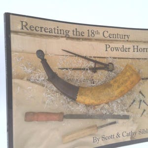 May include: A framed display featuring a powder horn and tools, with the text "Recreating the 18th Century Powder Horns." The horn is tan and brown, with a dark brown end. Tools include a file and a chisel.