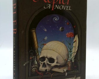 May include: A hardcover book titled "Kepler A Novel" by John Banville. The cover features a skull with a celestial globe, a telescope, and a feather, set against a dark background with the title in red script.