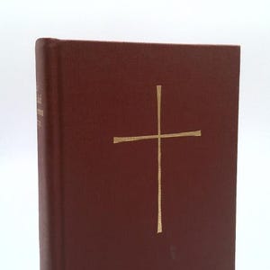 May include: A maroon hardcover book with a textured fabric cover. A thin, gold-colored cross is centered on the front cover. The spine of the book features gold-colored text that is partially visible and a small gold-colored emblem at the bottom.
