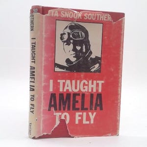 May include: A hardcover book titled I Taught Amelia to Fly by Neta Snook Southern. The red dust jacket features a black and white illustration of a pilot wearing goggles and a leather cap. The cover shows significant wear with torn edges.