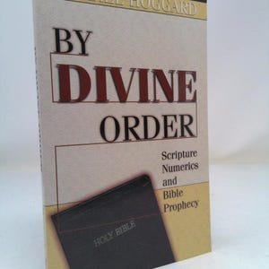 May include: A book titled "By Divine Order: Scripture Numerics and Bible Prophecy" by Michael Hoggard. The cover features a closed black Bible with the words "Holy Bible" embossed on the front.