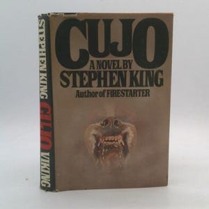May include: A hardcover book titled "CUJO" by Stephen King. The cover is brown with the title in large black letters and an illustration of a snarling dog. The spine reads "CUJO" and "Stephen King".