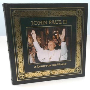 May include: A hardcover book with a black cover and gold accents. The book features a photograph of John Paul II with his arms raised. The title "JOHN PAUL II" is displayed at the top, with "A LIGHT FOR THE WORLD" at the bottom.