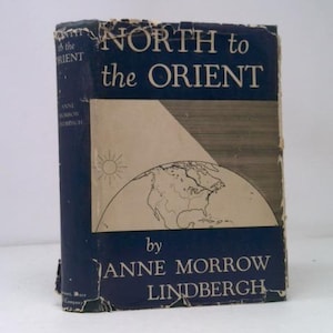May include: A vintage book titled "NORTH to the ORIENT" by Anne Morrow Lindbergh. The cover is a dark blue with white text and a graphic of a sun, a globe, and a stylized airplane. The book shows signs of wear.