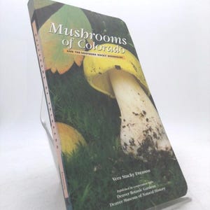 May include: A hardcover book titled "Mushrooms of Colorado" with a photograph of mushrooms on the cover. The book is propped up on a clear plastic stand. The author's name is Vera Stucky Evenson.