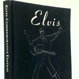 May include: Black commemorative book titled "Elvis" with a white illustration of Elvis Presley playing a guitar. The book's spine reads "Elvis-Commemorative Edition". A guitar-shaped zipper pull is attached.