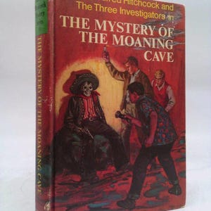 May include: Vintage hardcover book titled "The Mystery of the Moaning Cave" by Alfred Hitchcock and The Three Investigators. The cover features an illustration of a skeleton in a hat and three young men with flashlights. The spine reads "The Mystery of the Moaning Cave".
