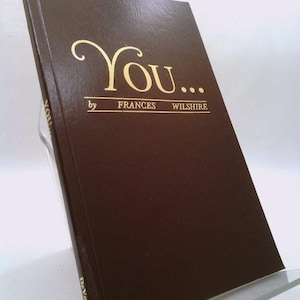 May include: A hardcover book titled "You..." by Frances Wilshire. The book has a dark brown cover with gold lettering. The book is propped up on a clear stand.
