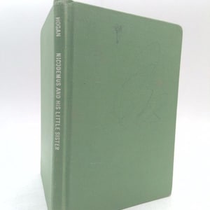 May include: A hardcover book with a light green cloth cover. The spine features the text HOGAN and NICODEMUS AND HIS LITTLE SISTER in white lettering. The front cover has faint pencil markings and a small, light-colored circular mark near the top.