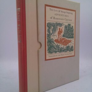 May include: A hardcover book titled "The Life & Strange Surprising Adventures of Robinson Crusoe" in a slipcase. The book's spine is red, and the slipcase is off-white with an illustration of Robinson Crusoe on a raft.