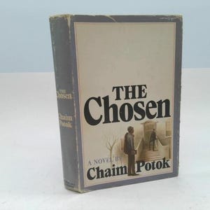 May include: A hardcover book titled "The Chosen" by Chaim Potok. The cover features a beige background with the title in large black letters and an illustration of two figures. The book has a blue spine and edges.