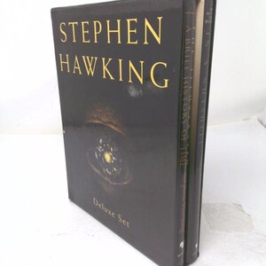 May include: A black box set containing two books by Stephen Hawking. The front cover features the author's name in gold lettering, the text Deluxe Set, and a central graphic of a glowing celestial object. The spine shows the titles A Brief History of Time.