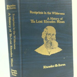 May include: A hardcover book titled "Footprints in the Wilderness: A History of The Lost Rhoades Mines." The cover is dark blue with gold lettering and a gold illustration of a man. The spine reads "A History of The Lost Rhoades Mines" and "Dream Garden Press."