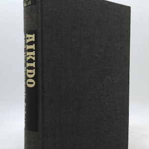 May include: A hardcover book titled "Aikido" with a dark gray textured cover. The title is vertically printed in gold lettering on a black spine. The book's spine also includes the publisher's name.