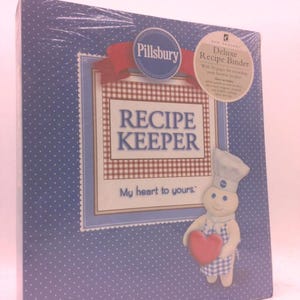 May include: A blue recipe binder with white polka dots, featuring the Pillsbury logo and the words "Recipe Keeper." A cartoon character holds a red heart. The binder includes a red ribbon and a sticker that says "Deluxe Recipe Binder."