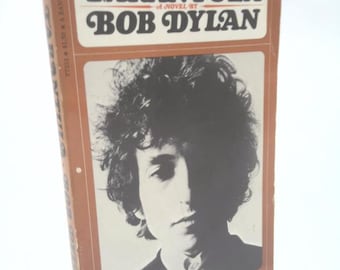 May include: A vintage paperback book titled "Tarantula" by Bob Dylan. The cover features a black and white portrait of Dylan. The book's title and author's name are in bold, retro-style lettering. The book is a Bantam Book.