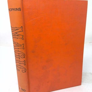 May include: A hardcover book with a bright orange cover. The spine features the black text "HOPKINS" at the top, the word "MAGIC" written vertically in a stylized black font in the center, and the word "ARNO" printed in black at the bottom.
