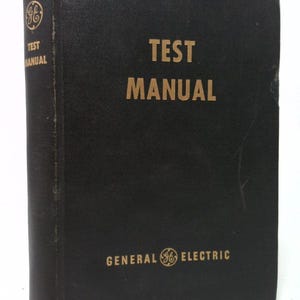 May include: A black "Test Manual" book with gold lettering and the General Electric logo. The book's spine also features the text "Test Manual" and the GE logo. The book appears to be vintage.