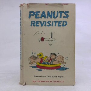 May include: A hardcover book titled Peanuts Revisited, Favorites Old and New by Charles M. Schulz. The off-white cover features blue text and a cartoon illustration of characters in a red and yellow inflatable pool with a dog jumping above them.