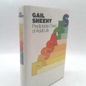 May include: A hardcover book titled "Predictable Crises of Adult Life" by Gail Sheehy. The book's cover is white with the title in black and the author's name in a larger font. The word "PASSAGES" is in colorful, stacked letters.