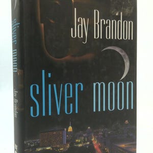 May include: A hardcover book titled "Sliver Moon" by Jay Brandon. The cover features a dark background with a cityscape at the bottom, a crescent moon, and the title in blue and white lettering. The author's name is in white.