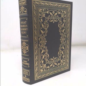 May include: A black leather-bound book with gold lettering and a decorative gold border. The title of the book is "What Makes Sammy Run?" by Budd Schulberg. The book is a signed edition.