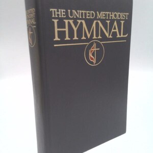 May include: A black hardcover book titled The United Methodist Hymnal with gold lettering on the front cover and spine. The cover features a circular emblem containing a cross and a flame symbol in gold and red, positioned below the title text.