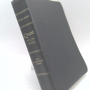 May include: A black hardcover Holy Bible, Quest Study Bible, New International Version, published by Zondervan. The spine features gold-colored text and horizontal lines, with a textured cover surface and a black ribbon bookmark extending from the bottom.