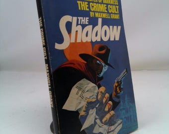 May include: Vintage paperback book titled "The Shadow" by Maxwell Grant. The cover features a masked figure in a hat holding a gun, set against a dark blue background with an orange circle. The book's title is in large white letters.