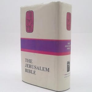 May include: A hardcover edition of "The Jerusalem Bible" with a cream-colored cover. The book features a purple and pink band across the middle and decorative elements in pink. The title is printed in black.