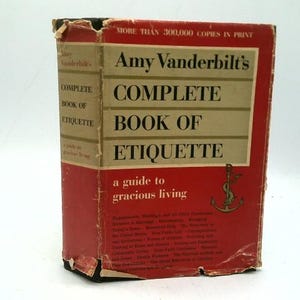 May include: A vintage copy of "Amy Vanderbilt's Complete Book of Etiquette." The book has a red cover with gold lettering and a gold anchor emblem. The title and author's name are prominently displayed.