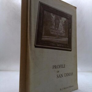 May include: A vintage book titled "Profile of San Dimas" by J. Howard Hoover. The cover features a sepia-toned image of a house within a dark frame. The book has a worn, aged appearance with a light beige cover.
