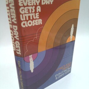 May include: A hardcover book titled "Every Day Gets a Little Closer." The cover features a design with orange, brown, and blue hues, with the title in white block letters. The author's names are also visible.