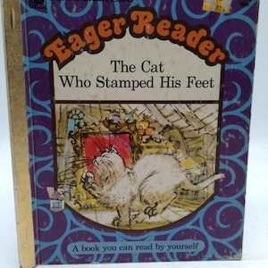 May include: Vintage children's book titled "Eager Reader: The Cat Who Stamped His Feet." The cover features a white cat illustration inside a framed image. The book has a purple and blue design with gold spine.