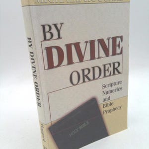 May include: A book titled "By Divine Order" by Michael Hoggard. The book cover features a closed Bible with the words "Holy Bible" embossed on the front. The book cover also includes the text "Scripture Numerics and Bible Prophecy".