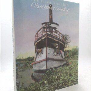 May include: A book titled "Okeechobee County" with a vintage image of a boat on the cover. The boat is white with the word "SUCCESS" on the front. The book's title is in a cursive font.