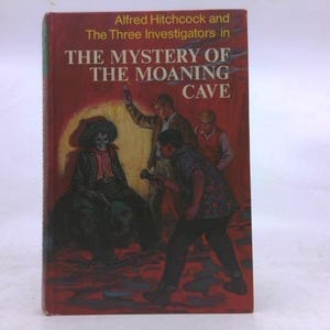 May include: A vintage book titled "The Mystery of the Moaning Cave" by Alfred Hitchcock. The cover features an illustration of three men and a skeleton in a hat. The background is a dark red with yellow highlights.