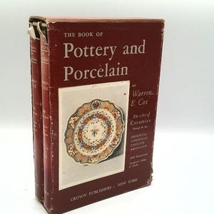 May include: A vintage hardcover book titled "The Book of Pottery and Porcelain" by Warren E. Cox. The cover is a deep burgundy color with white text and an image of a decorative plate. The book is a guide to ceramics.