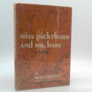 May include: A vintage hardcover book titled "Miss Pickthorn and Mr. Hare: A Fable" by May Sarton. The cover is a warm orange with an illustration and text in white. The book is standing upright.
