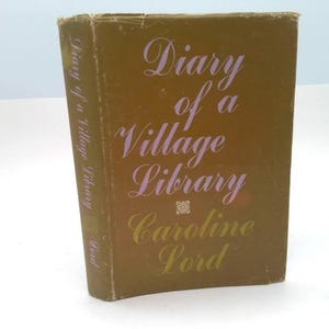 May include: A hardcover book titled "Diary of a Village Library" by Caroline Lord. The book has a brown cover with the title and author's name in a light purple and gold script font. The book is standing upright.