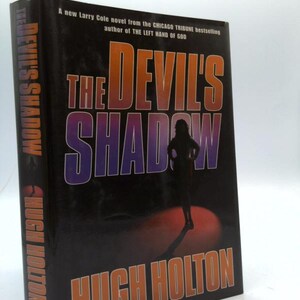 May include: A hardcover book titled "The Devil's Shadow" by Hugh Holton. The cover is black with orange and purple lettering. A silhouette of a person stands in a spotlight. Text on the spine reads "The Devil's Shadow" and "Hugh Holton."