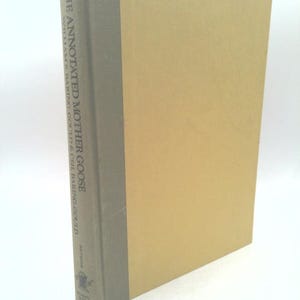 May include: A hardcover book titled The Annotated Mother Goose by William S. Baring-Gould and Ceil Baring-Gould. The book features a tan front cover and a gray spine with the title and author names printed in a dark font, along with a small logo.